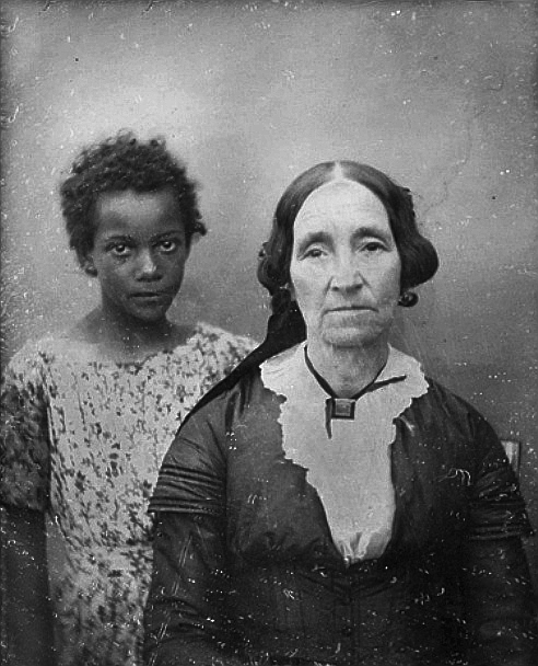 Daguerreotype portrait taken circa 1850 in New Orleans, Louisiana of an older white female with a young enslaved female servant. The older woman’s facial expression conveys a sense of tiredness and forlornness, and the young girl’s expression conveys a certain sadness, but there is a fire to her gaze as she look looks into the camera lens. 