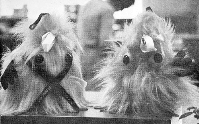 Black and white photograph of two West Springfield High School monsters, triangular stuffed animals covered with fuzzy fluff and two giant eyeballs, atop a library desk. 