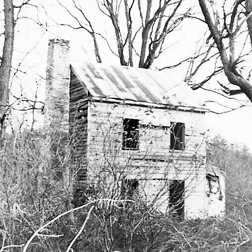 Black and white photograph of the Barker family home. It is two-stories tall and has a brick chimney on one side of the structure. The landscape around the home is overgrown with weeds and brambles.
